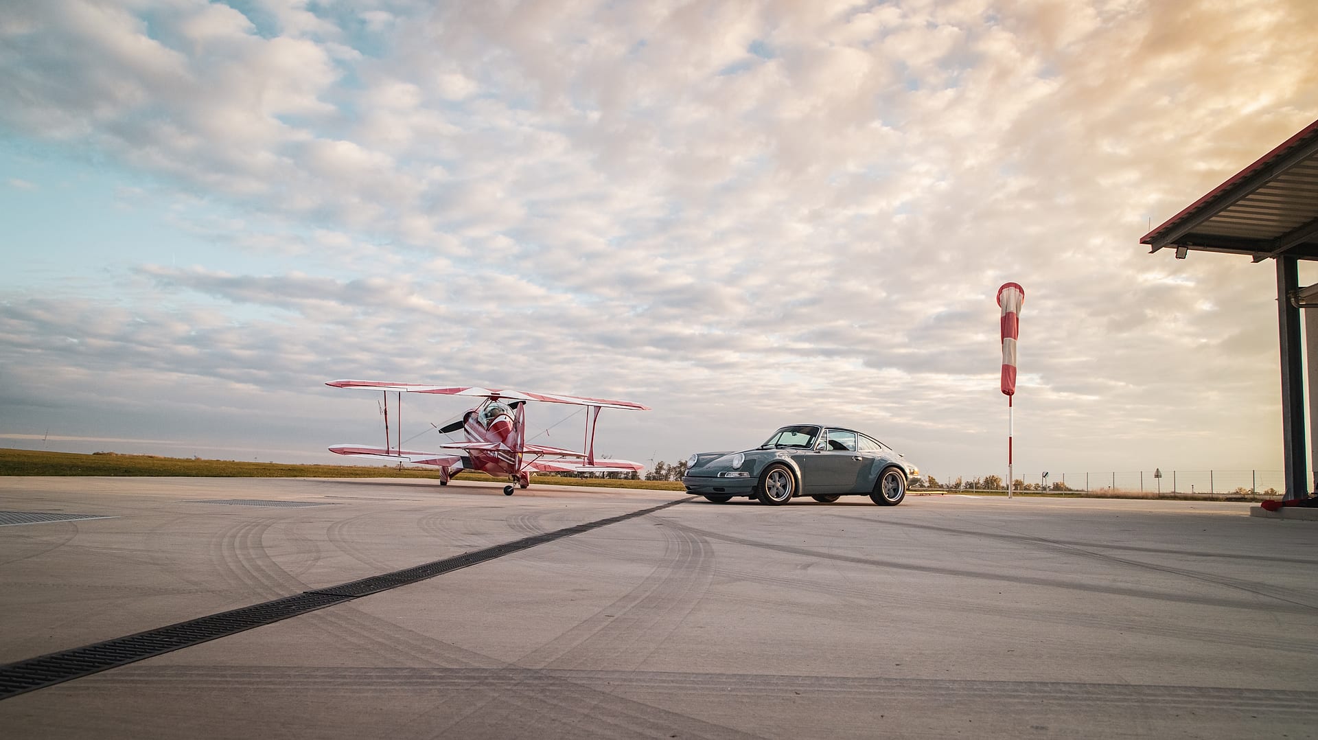 Klassischer Porsche 911 neben einem Doppeldecker auf einem Flugfeld im Abendlicht